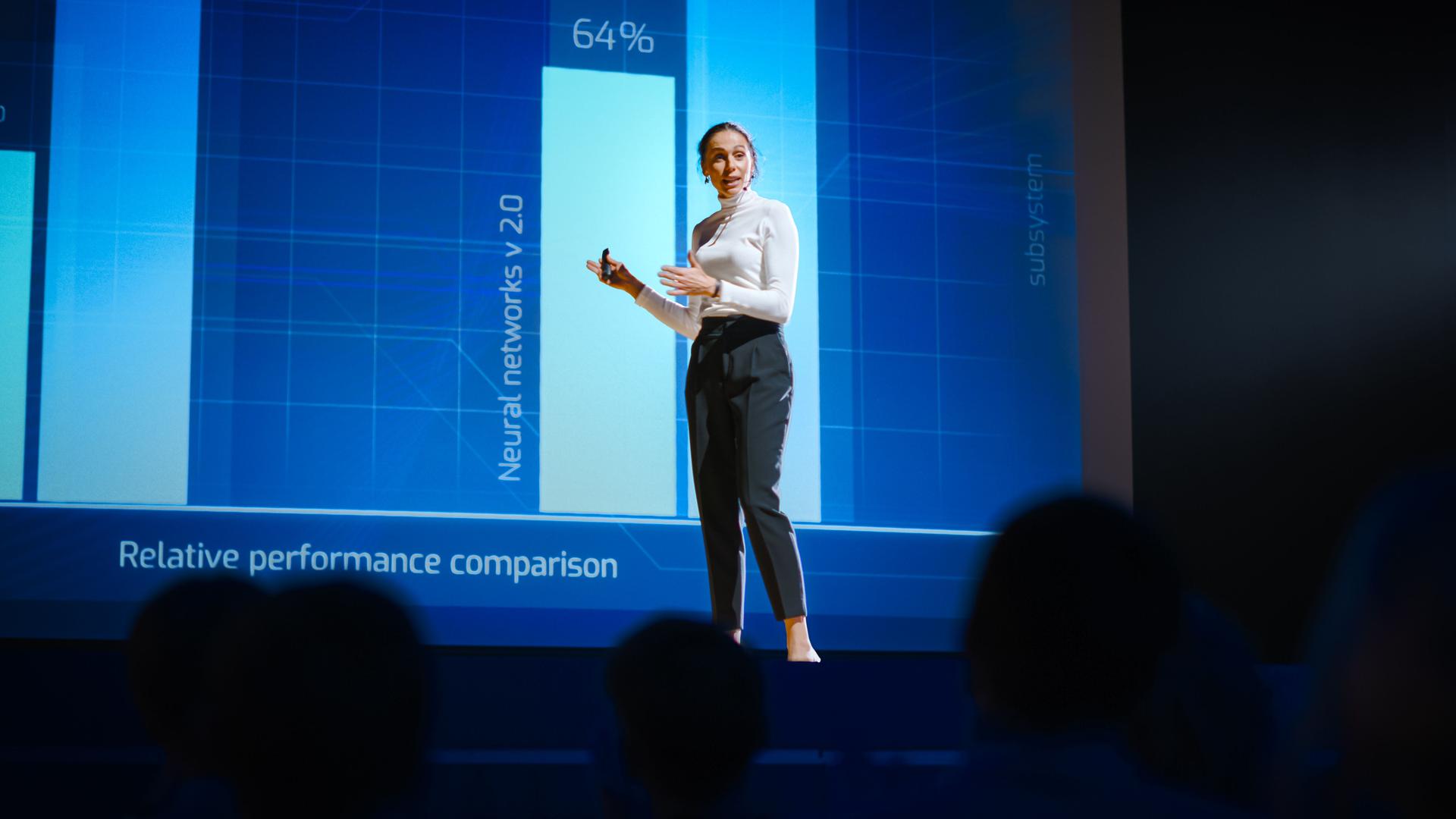 On Stage, Successful Female Speaker Presents Technological Product, Uses Remote Control for Presentation, Showing Infographics, Statistics Animation on Screen. Live Event / Device Release.